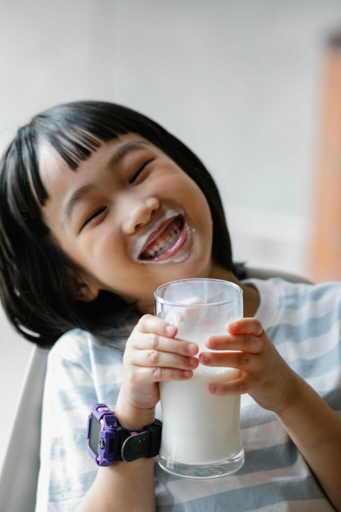 Cute little girl with smartwatch toothy smiling while holding glass of milk in hands